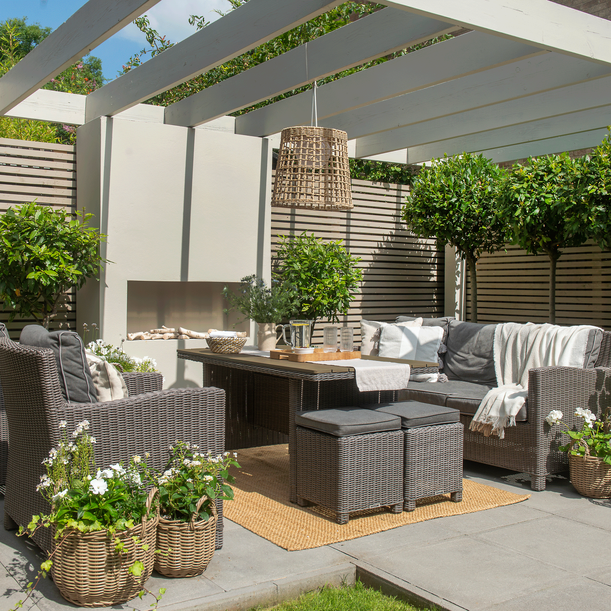a compact garden with patio with a large pergola overheaf horizontal slatted fencing a table and sofa seating an open outdoor fireplace and bay trees