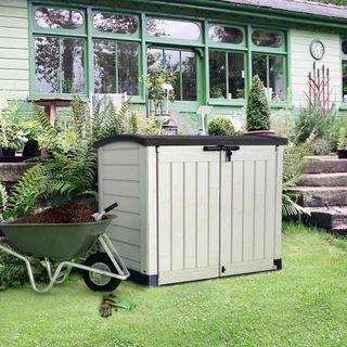 Grey storage unit with charcoal lid and 2 doors standing on green grass next to wheelbarrow with garden soil inside