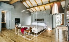 A hotel bedroom with a four post bed, cow skin chairs, a desk, a fireplace, a floor lamp, wooden floors and wooden ceilings.