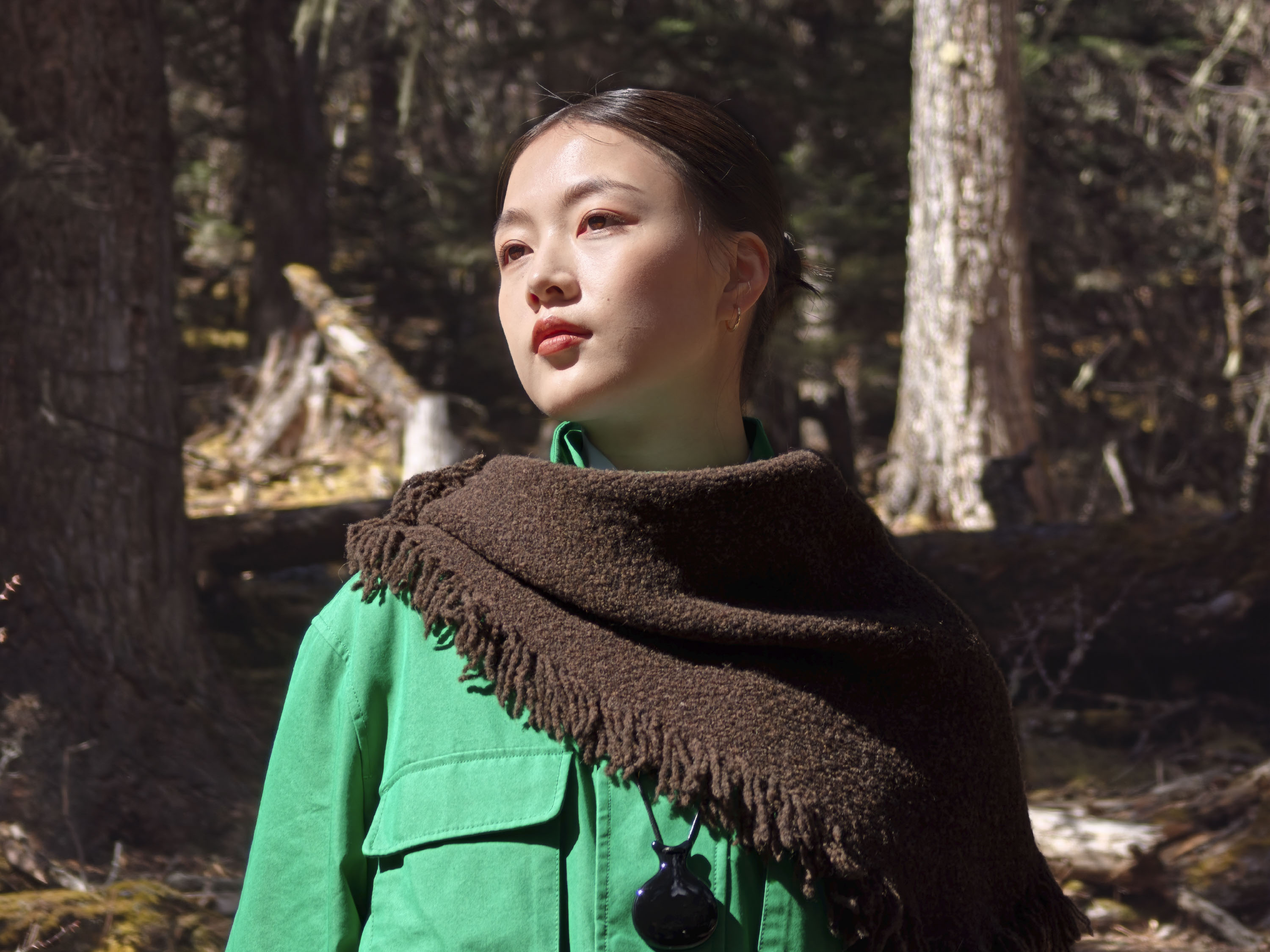 Portrait of a woman in a shawl and green jacket in a woodland, there's direct sunlight on her