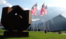 Daytime, outside image of Interior 08, Kortrijk Belgium, green grass, large brown sculpture in forefront, white marque tent, white flag poles with flying flags, visitors, blue cloudy sky