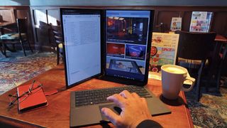 A dual-screen laptop is set up in a vertical orientation with the detachable keyboard in front, and a hand is shown resting on the trackpad.