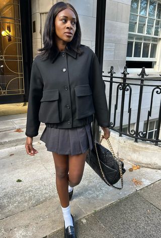 A woman wearing a gray pleated miniskirt with a black blouson jacket and black loafers.