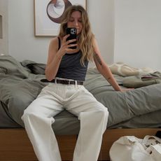 Influencer @brittanybathgate sits on a bed wearing white jeans with brown fisherman sandals and a black tank top.