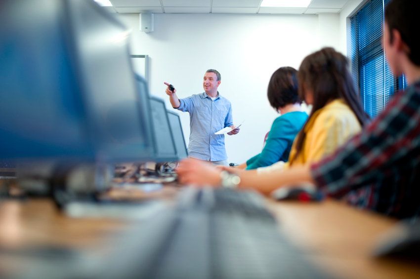 Kids at desktop computers listen to a male teacher as he speaks.