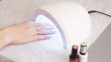 A woman's hand having a gel nail treatment, curing the nails under a UV lamp, with bottles of nail varnish beside 