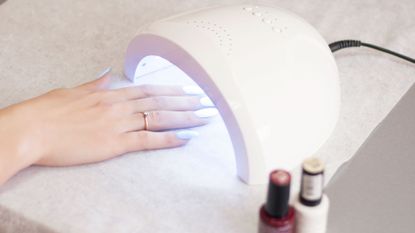 A woman's hand having a gel nail treatment, curing the nails under a UV lamp, with bottles of nail varnish beside