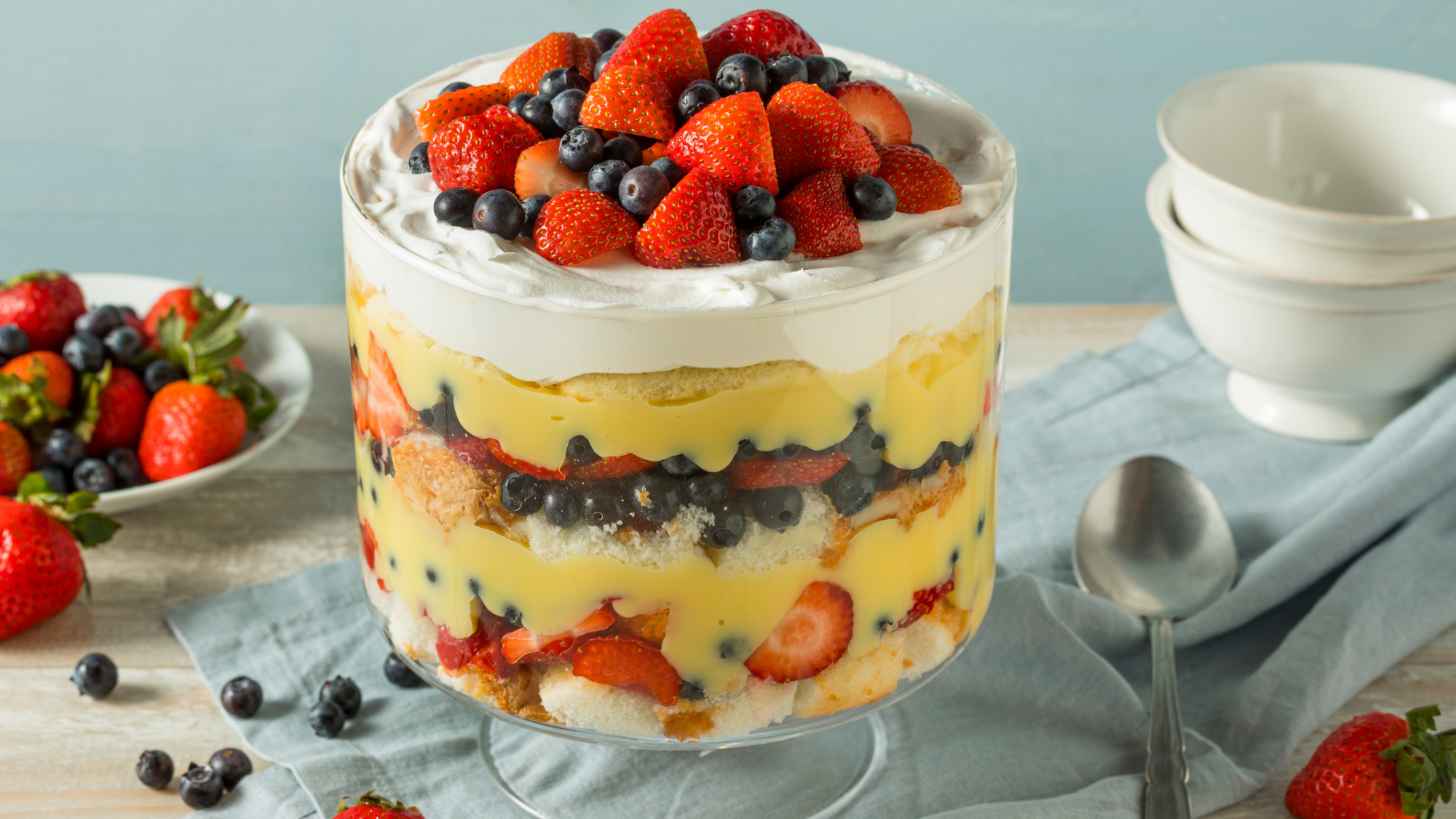 Trifle on a blue table cloth next to bowls of fruit