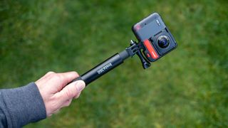 A hand holding an Insta360 selfie stick and camera in front of grass