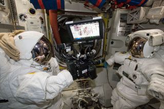 A RED Epic Dragon cinema camera floats between two spacesuits inside the Quest airlock on the International Space Station.