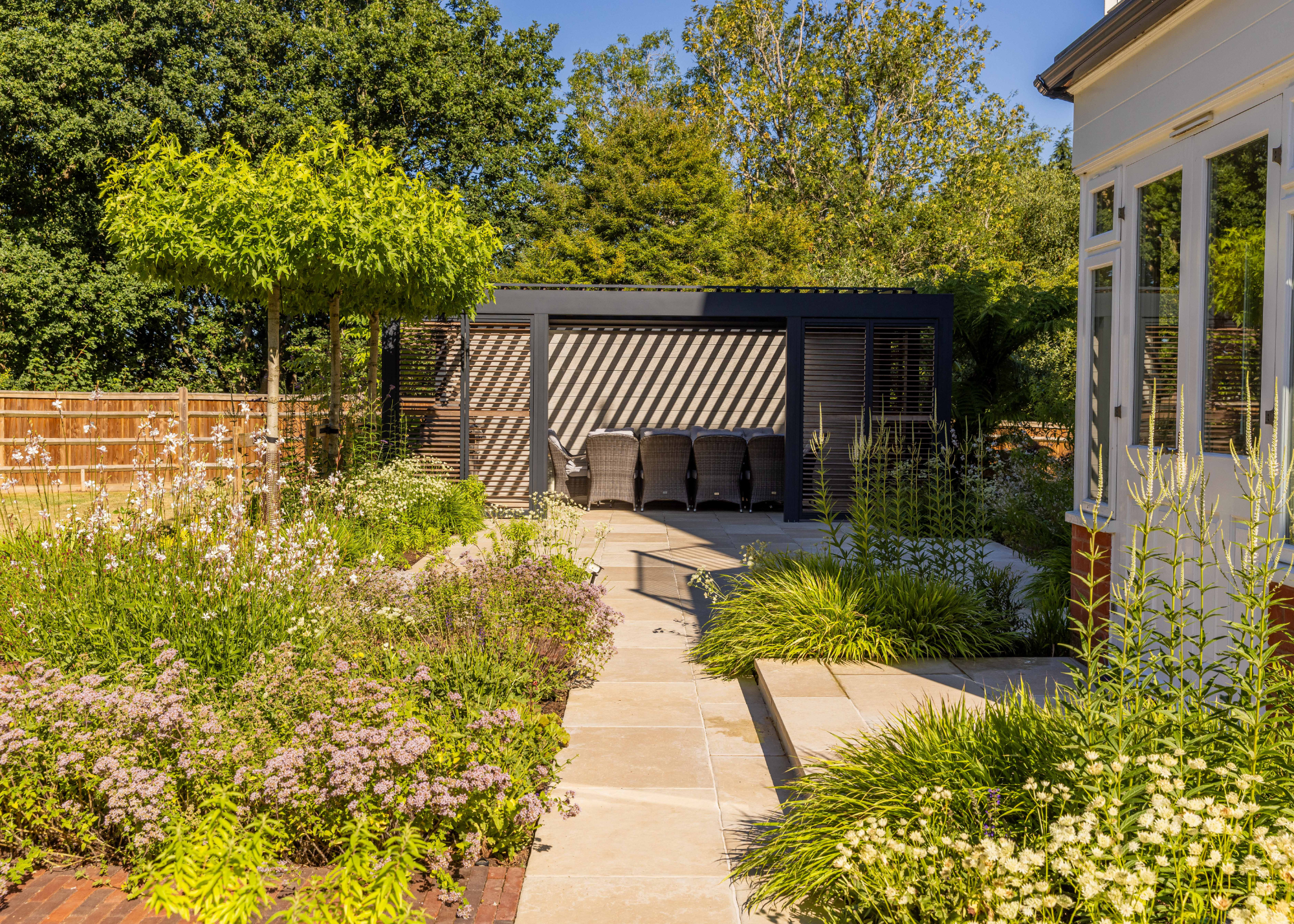 a sun-soaked garden with light-colored paving, beautiful shrubbery and a covered pergola