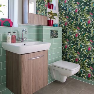 bathroom with botanical wallpaper and green tiles wall commode wash basin and wooden cabinet