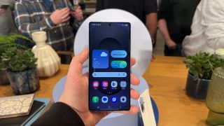 The Samsung Galaxy S25 Ultra being held in front of a table with people milling around in the background. The phone is showing its home screen.