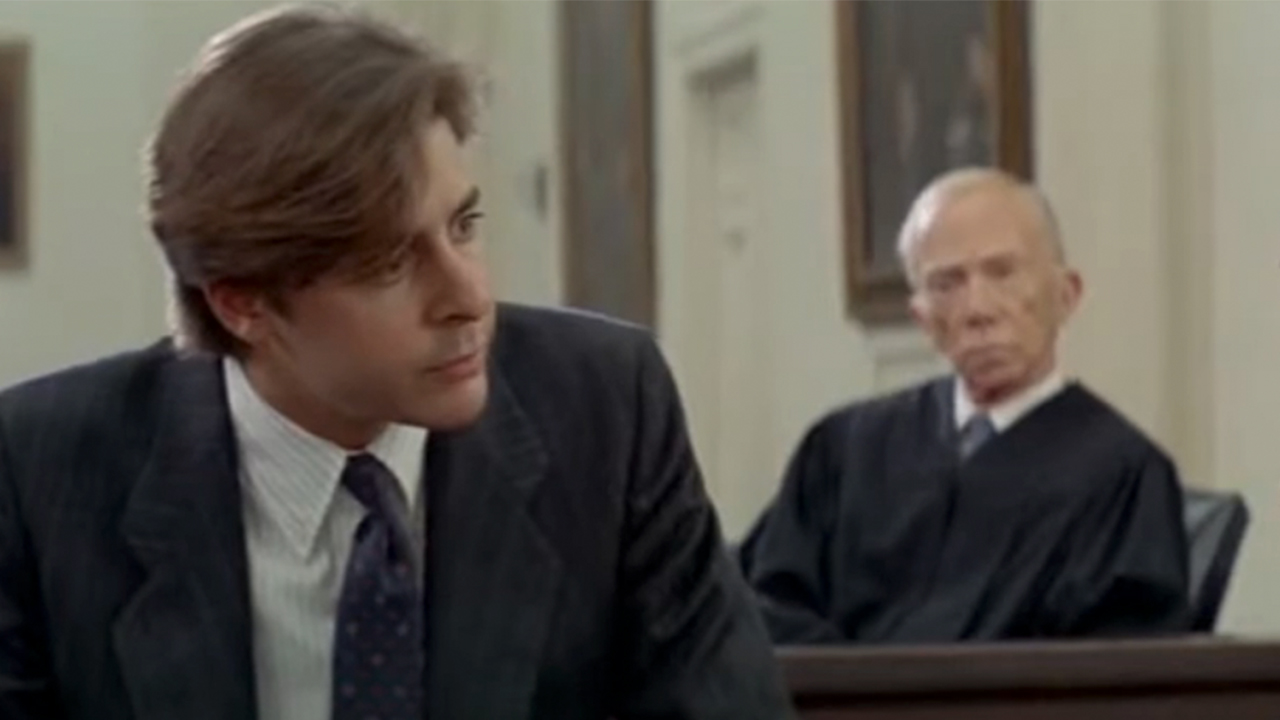 Judd Nelson in a suit looking to his left, in front of a judge played by Ray Walston in From The Hip