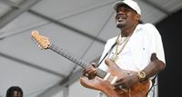 Eric Gales smiles (or grimaces?) as he hits a note and holds it onstage with his S-style signature guitar.