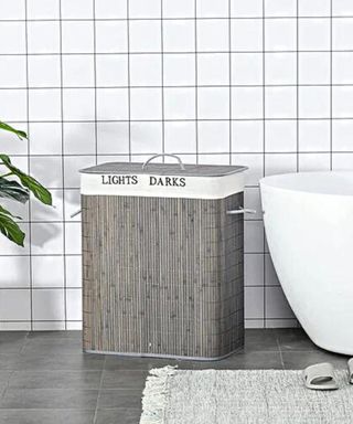 A grey split laundry hamper in a white tiled bathroom