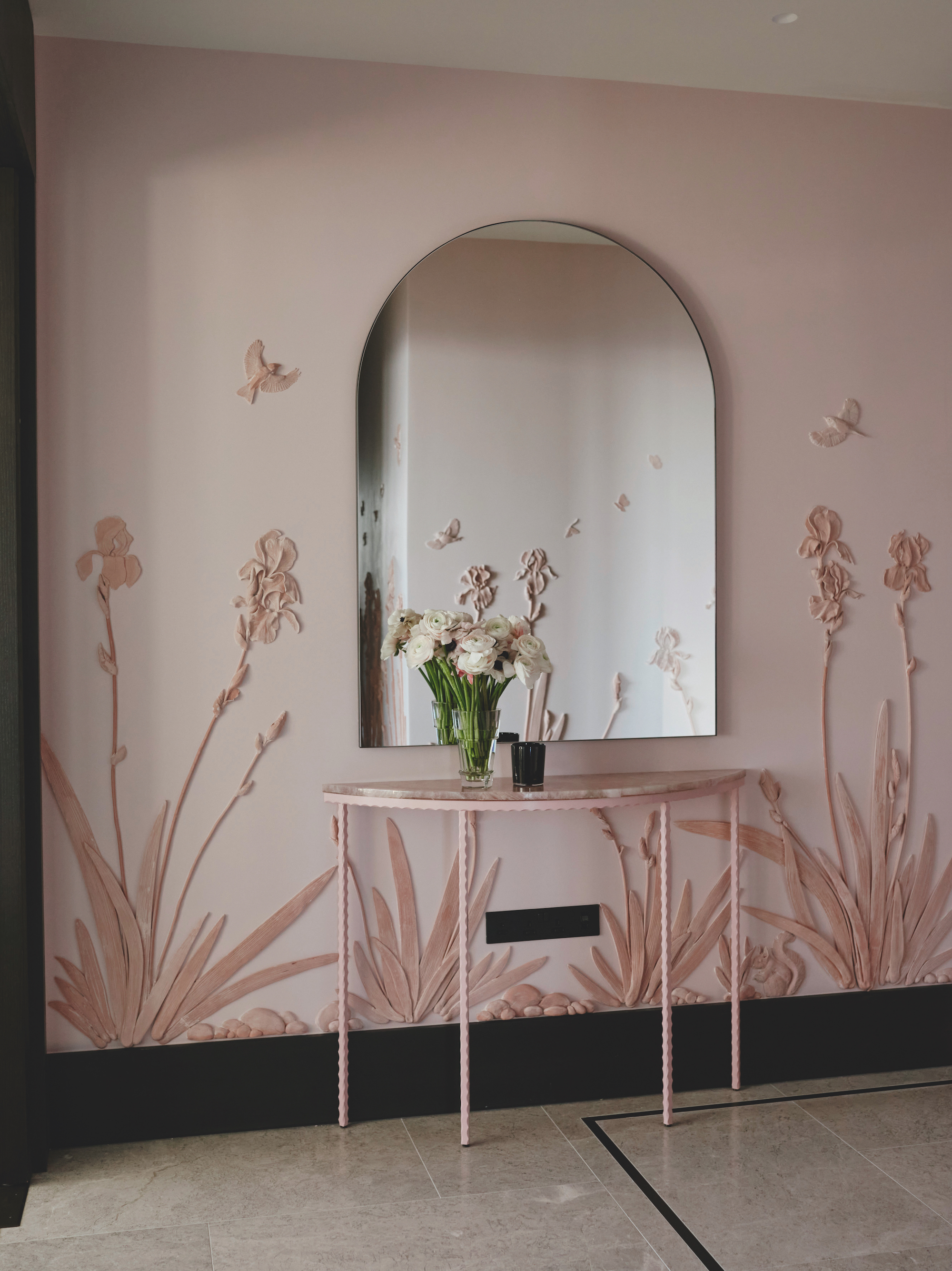 A floral plaster design in an entryway, painted in a soft blush pink, with a curved mirror above a pink entry table.