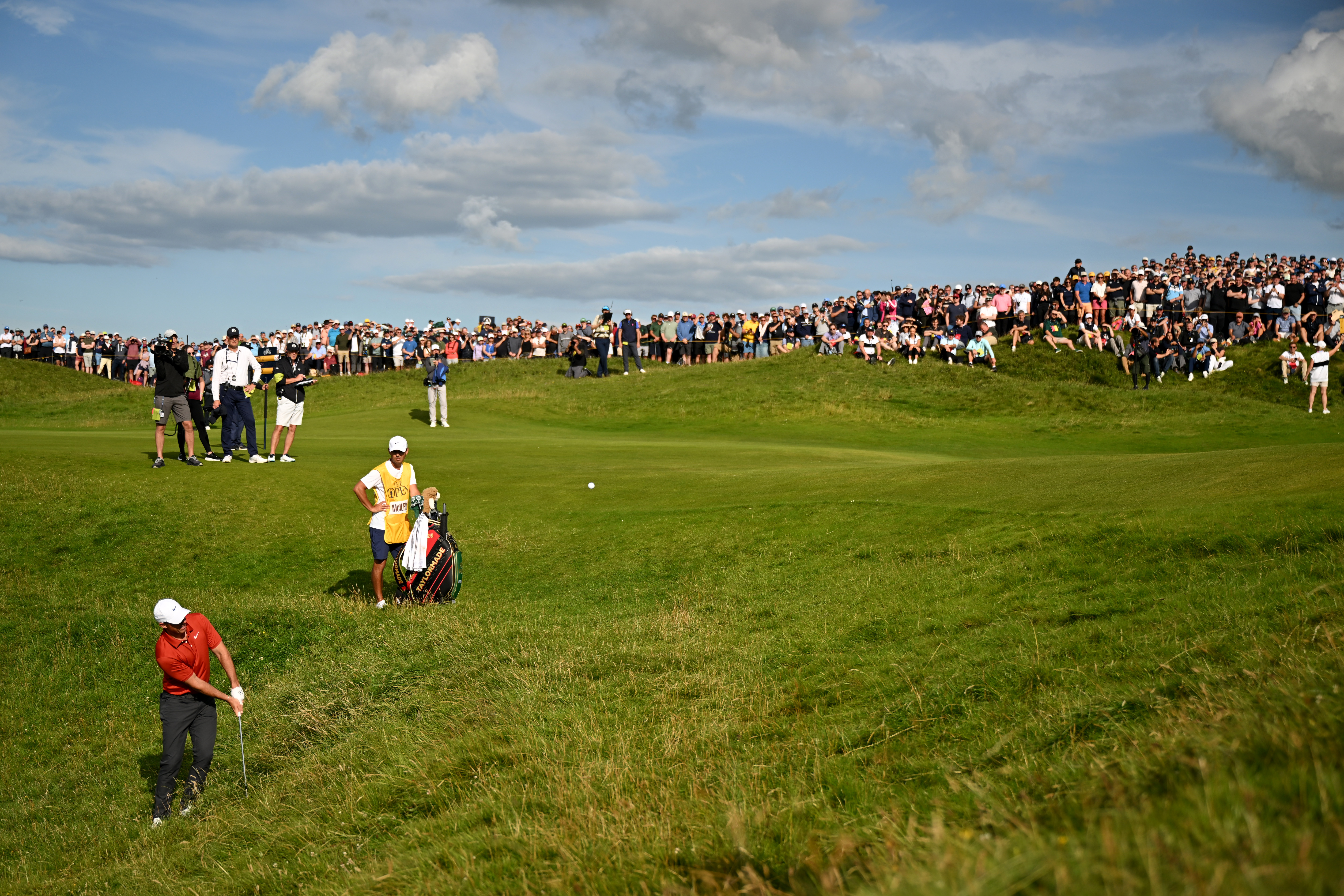 Rory McIlroy plays a chip shot (foreground) while huge galleries of fans and journalists watch on in the background