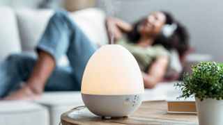 An oil diffuser diffusing with a woman laying on a couch and listening to music in the background