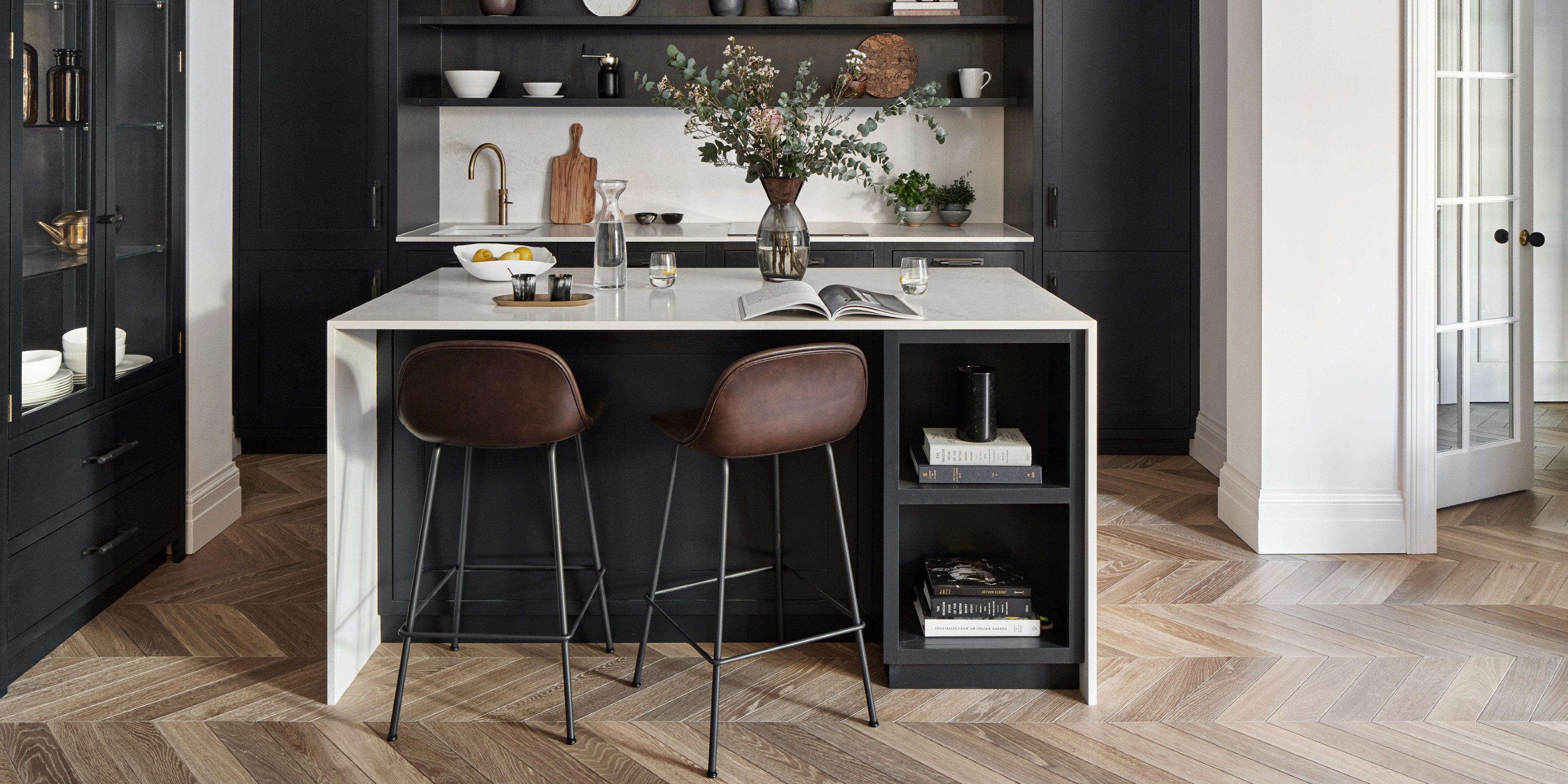 Dark kitchen cabinets with white worktops and wooden chevron floor, with brown leather bar stools against the island