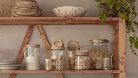 Glass food containers with dried food goods in them on a wooden shelving unit. 