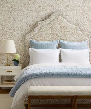 Blue and white bedding on an upholstered bed frame against white and gold patterned wallpaper; to the left, a lamp on a white nightstand.