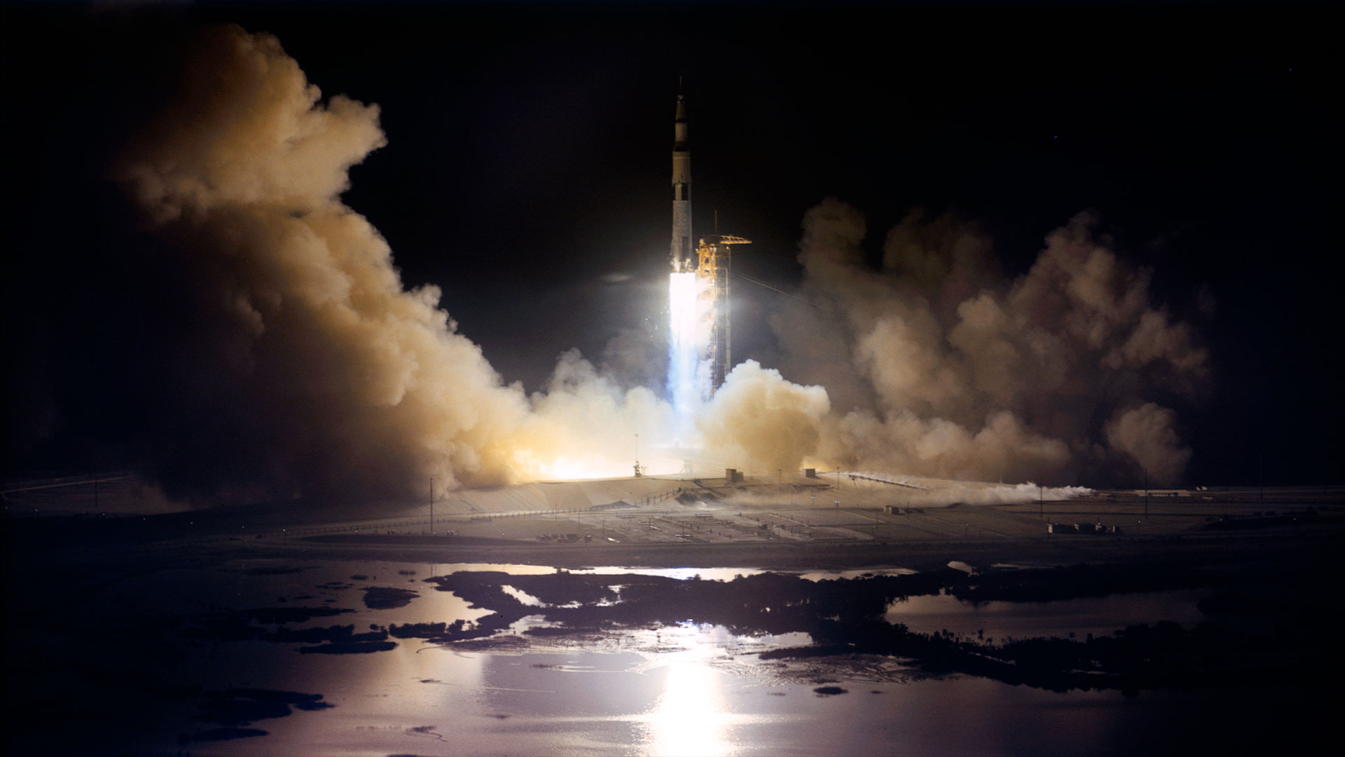 a large white rocket with black stripes lights up the horizon as it lifts off its launch pad at night