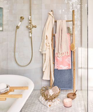 A bathroom with cream tiles, a gold shower, a wooden ladder shelf with pink and blue towels hanging from it, and a bath tub with a wooden tray on it