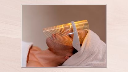 Side view of woman laying down under a towel and with one over her hair wearing a light therapy face mask