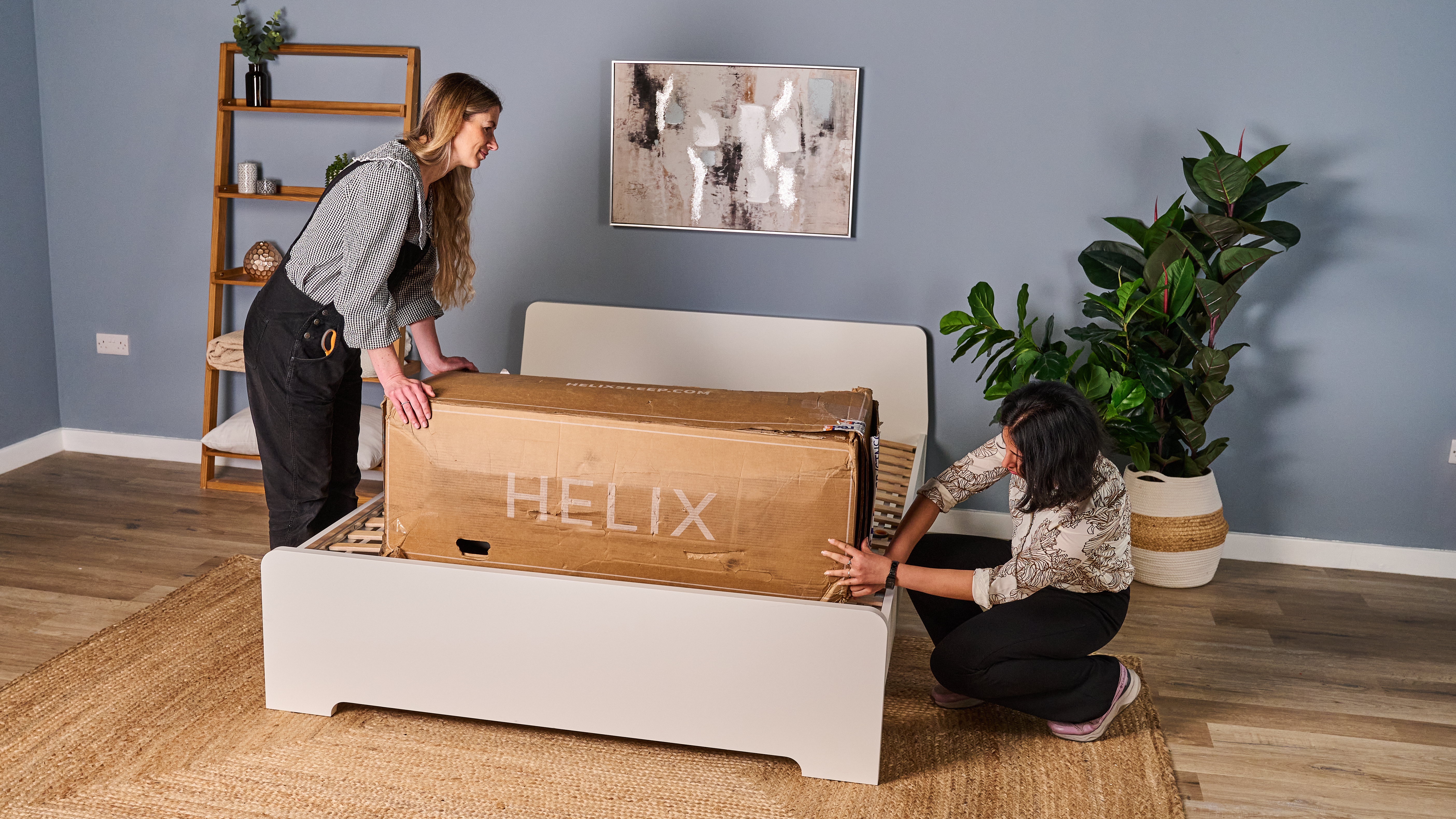 The image shows two mattress testers unboxing the Helix Midnight Luxe mattress and setting it up on a white bed frame in a blue bedroom