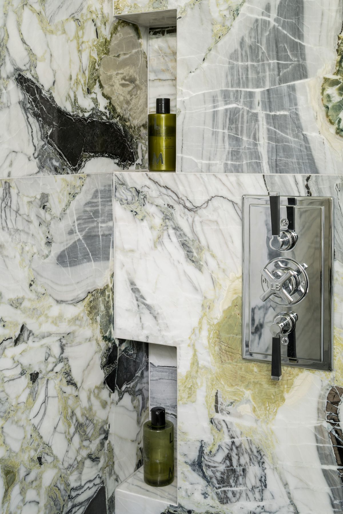 A shower tiled with green marble tiles, featuring a chrome shower fixture and green bottles of shampoo
