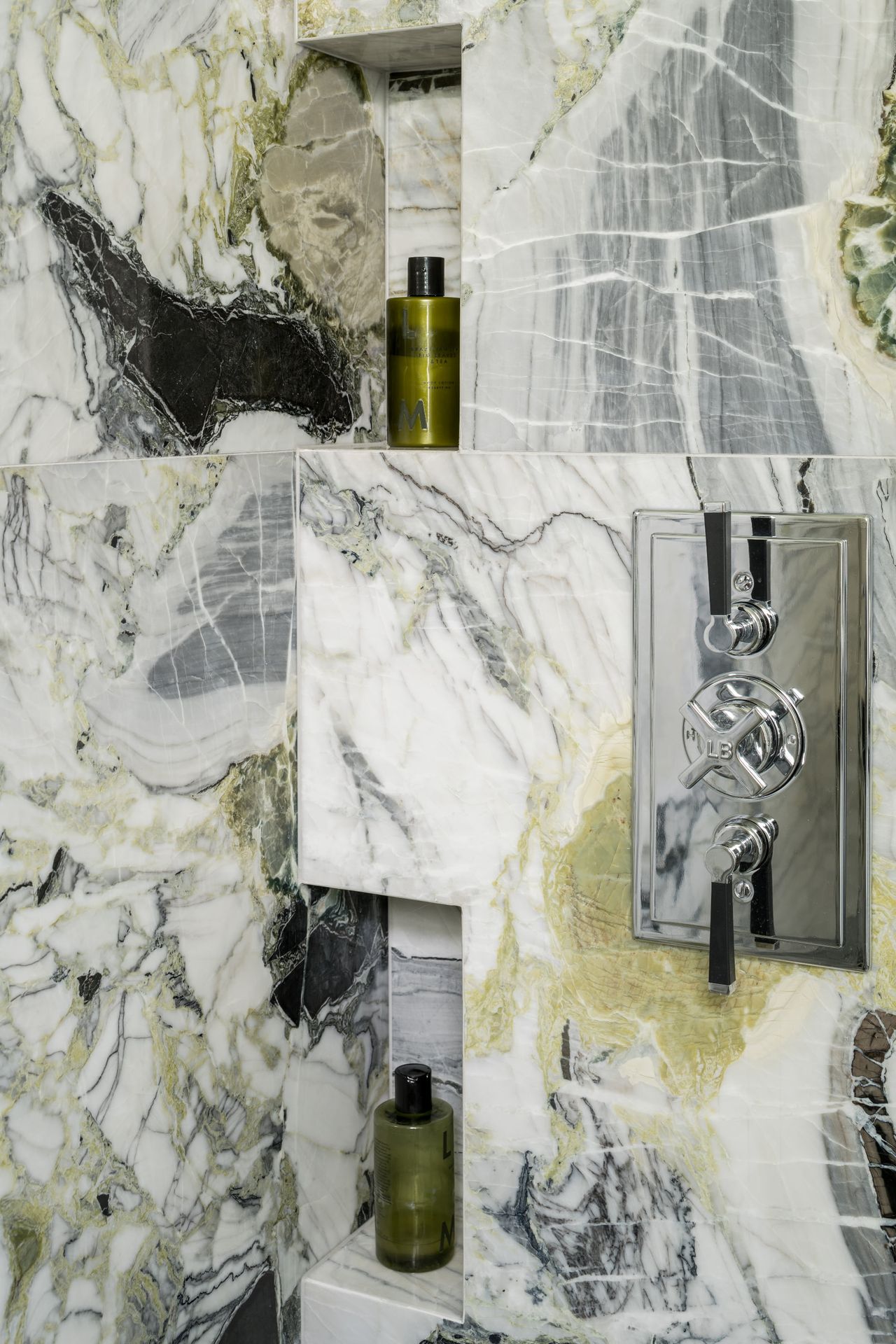 A shower tiled with green marble tiles, featuring a chrome shower fixture and green bottles of shampoo