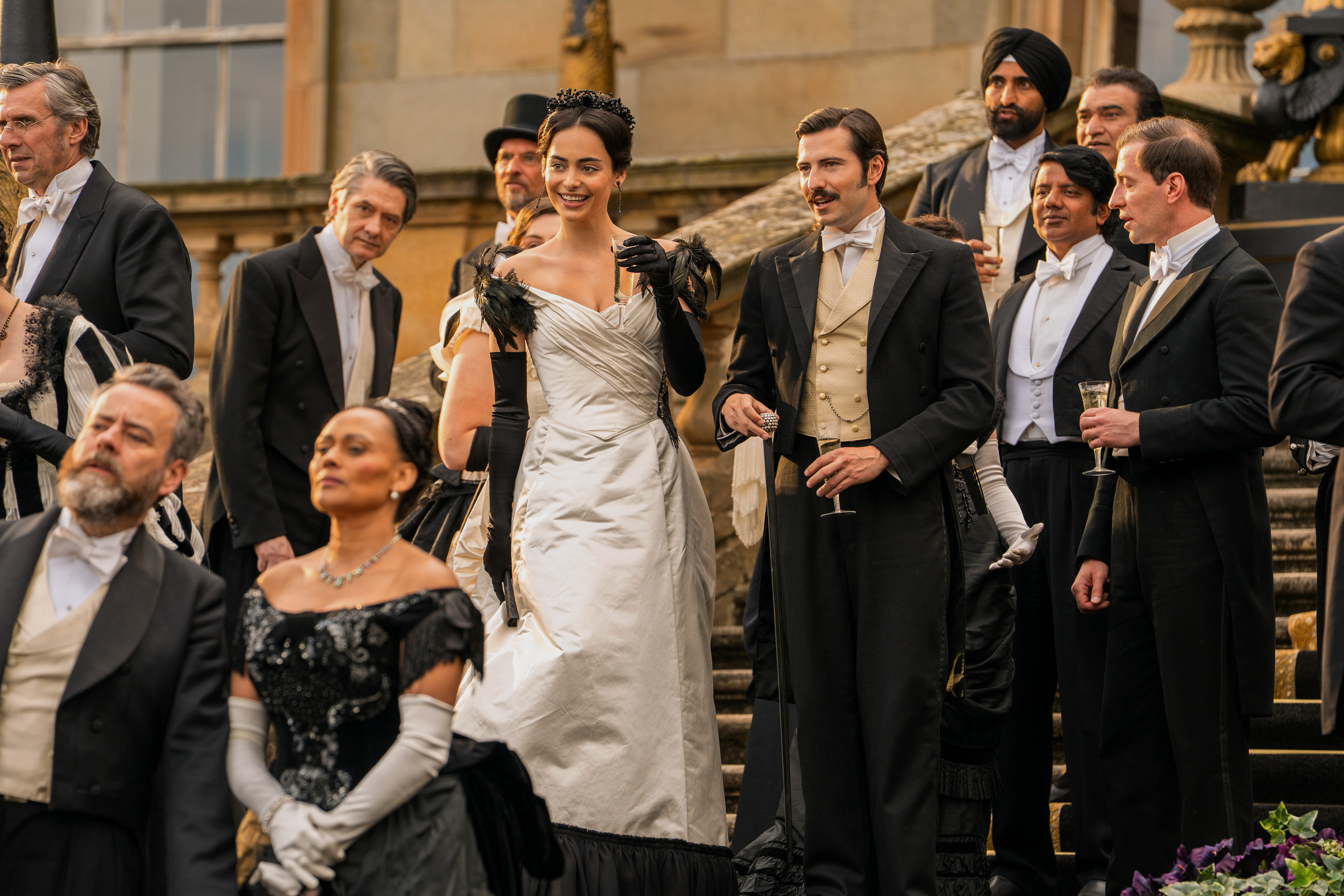 aubri Ibrag and Jacob Ifan wearing a silver dress and a tux standing in the center of a staircase surrounded by a crowd in the buccaneers season 2