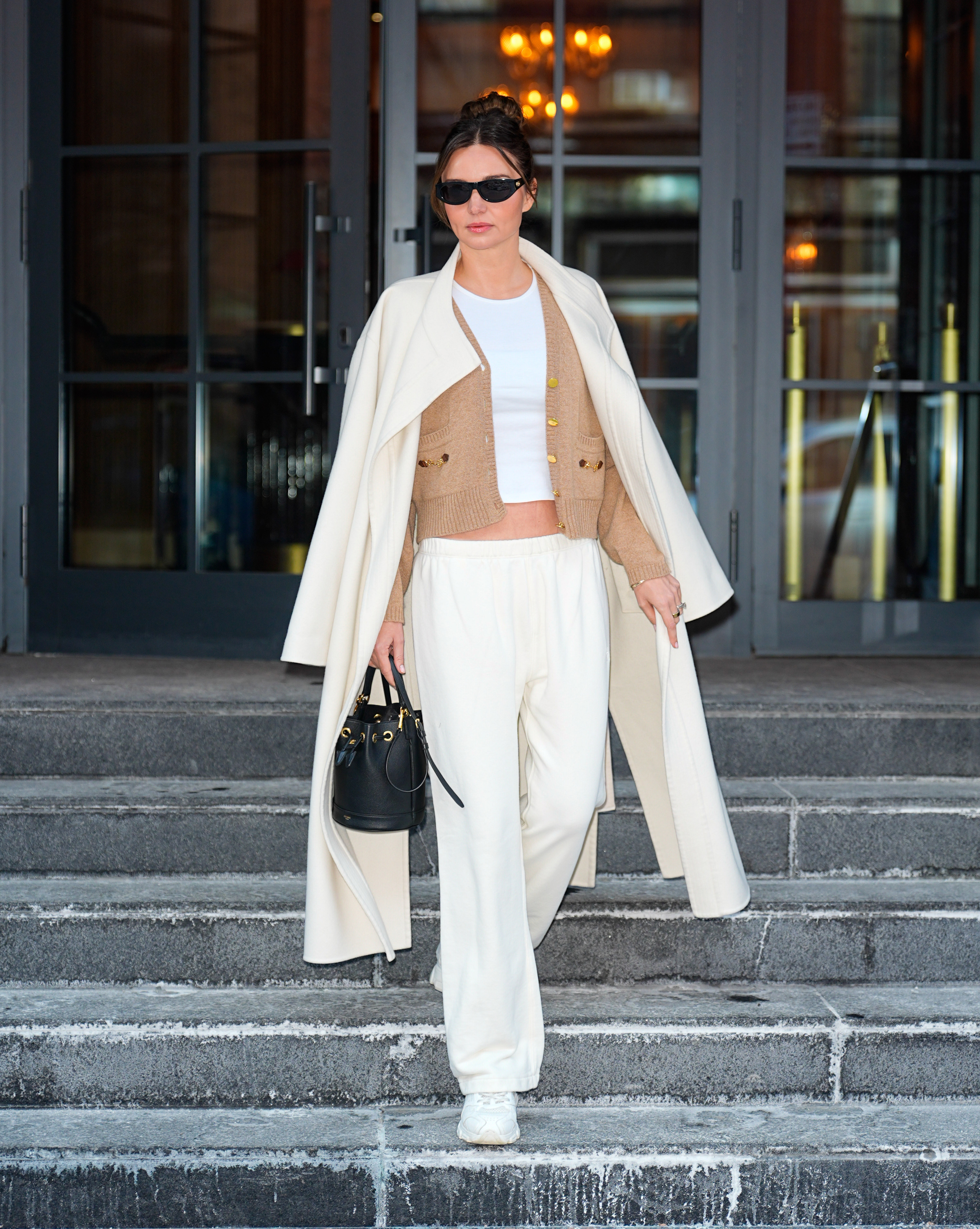 Miranda kerr wears an ivory coat, tan cardigan, white top, ivory pants, black bag, and white sneakers.