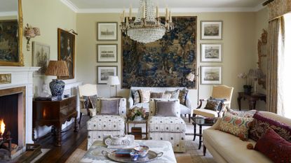 A formal, traditional living room featuring a grand crystal chandelier, a large blue landscape tapestry, and a crackling fireplace
