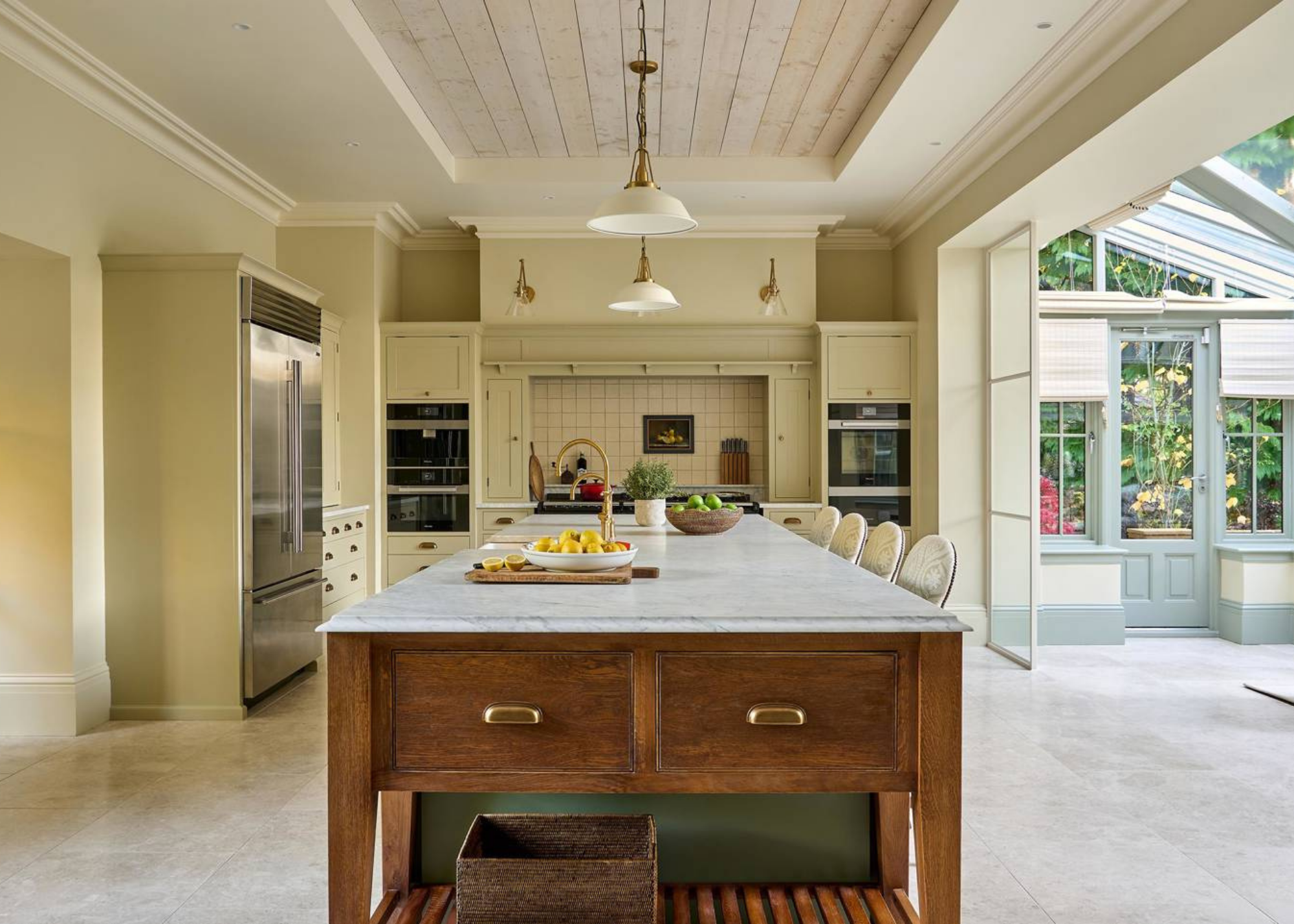 farmhouse style kitchen painted with butter yellow walls, a freestanding timber island with marble top, and a tiled area for the stove. There is an indoor conservatory next to the kitchen, panelled ceilings, and pendant lights hanging over the island