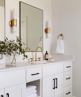 A contemporary white bathroom with brass faucet and light fittings, black cupboard handles and plants