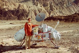 a man in a jacket stands beside a spacecraft in a desert