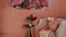 A dusty pink dressing room with a sofa seater, a plush pillow and a two-toned scarf, alongside a side table with a vase of flowers