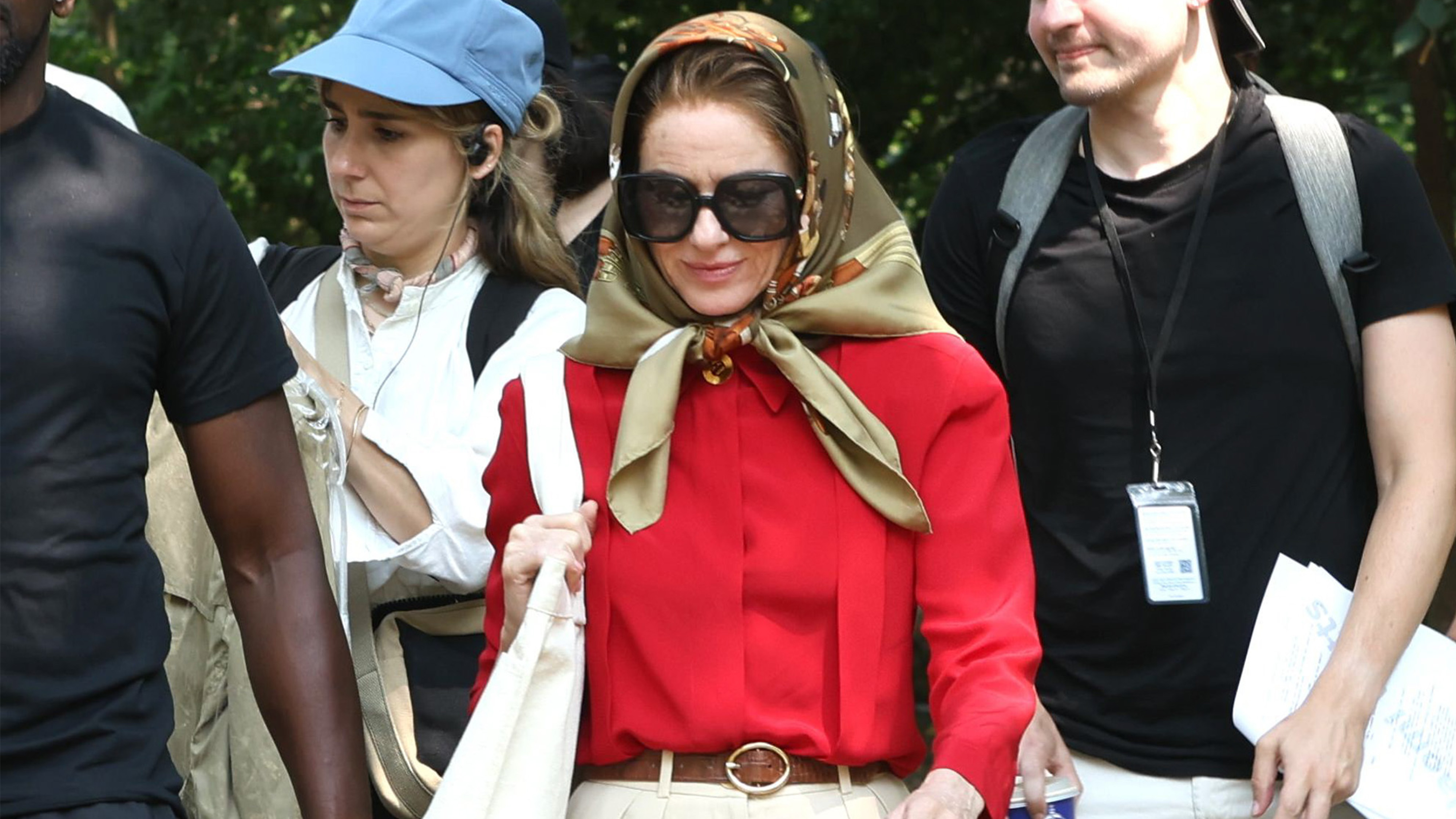 Naomi watts transforms into jackie kennedy wearing a headscarf, large sunglasses, a red blouse, brown belt, beige wide-leg pants, and brown woven leather ballet flats.