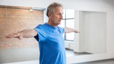 A man in a blue t-shirt stands in a gym studio with his arms out to his side. Behind him we see a mirror, reflecting a red brick wall and window.