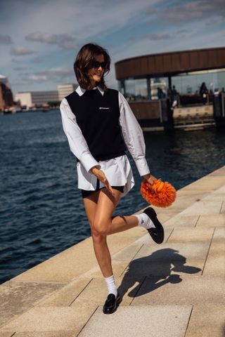 A woman at Copenhagen Fashion Week wearing a white button-down shirt, black sweater vest, white socks, and black loafers.