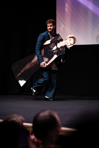 Andrew Garfield carrying a cardboard cutout of Florence Pugh on a dimly lit stage