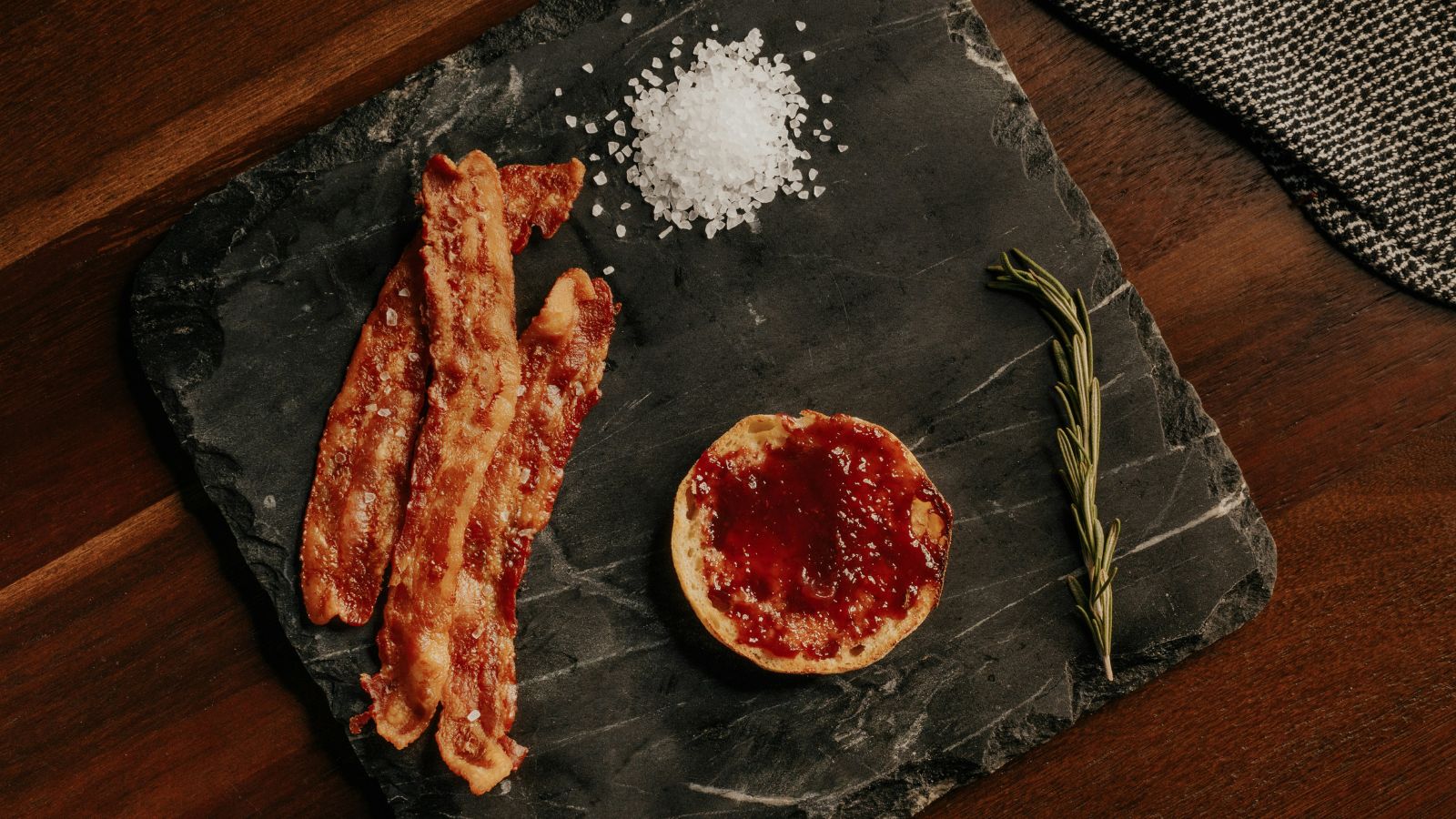 Three rashers of bacon on a slate tray, accompanied by a spring of rosemary, a small serving of salt, and a pot of red sauce