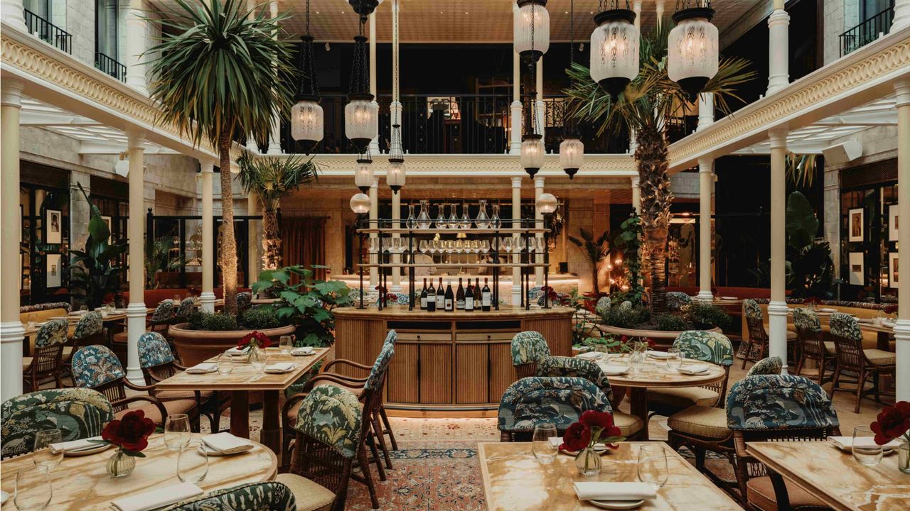 A restaurant dining room located in an indoor atrium filled with plants boasts layered interiors, including marble-topped tables, wood and textile upholstered armchairs with patterned fabric in blue and cream tones, intricate rugs in red tones, and glass and wrought iron pendant lights.