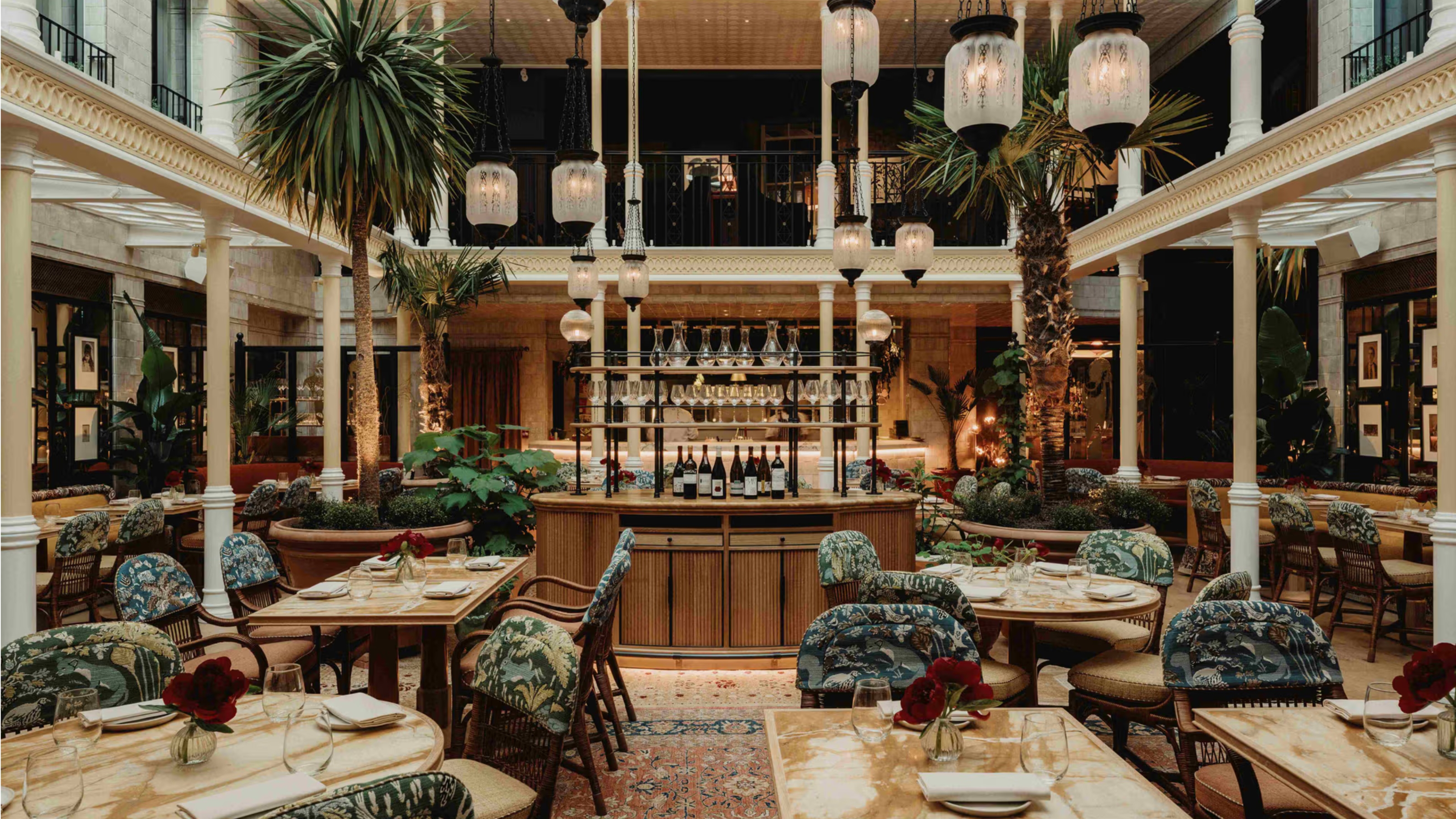 A restaurant dining room located in an indoor atrium filled with plants boasts layered interiors, including marble-topped tables, wood and textile upholstered armchairs with patterned fabric in blue and cream tones, intricate rugs in red tones, and glass and wrought iron pendant lights.