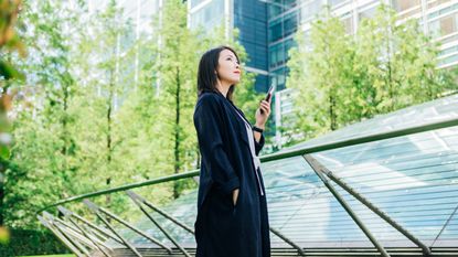 A young businesswoman on her smartphone stands outside an office building surrounded by greenery.