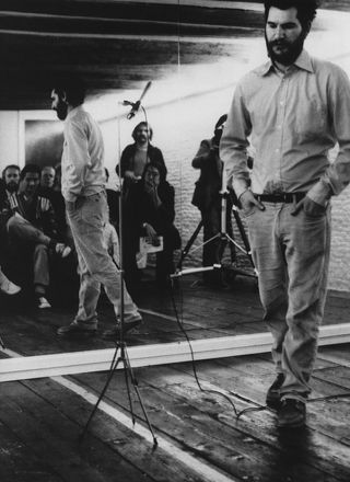 Black and white image, wooden floor, mirrored wall reflectinb the room of a seated small audience, a bearded male performer in shirt and jeans, with hands in pockets stood next to a microphone stand