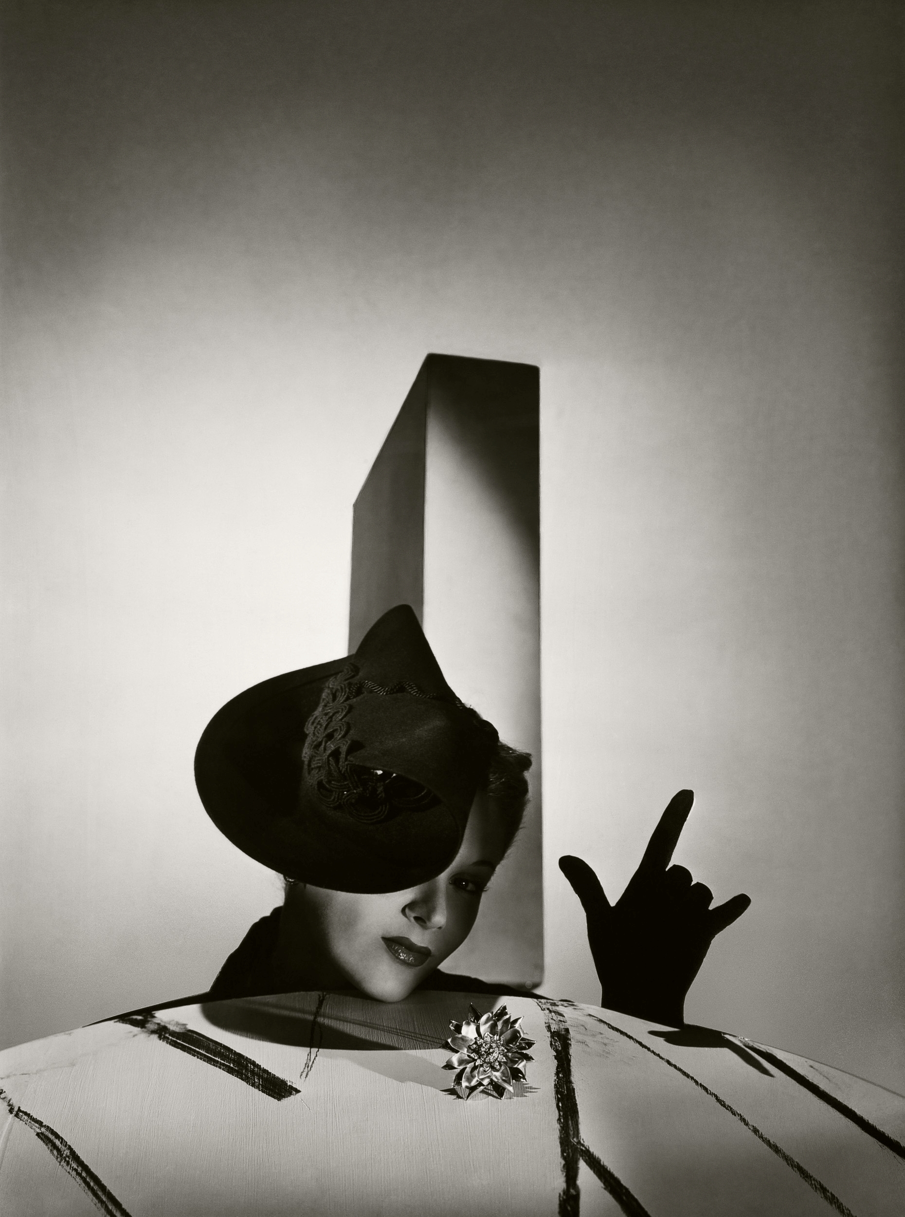 A model wearing an architectural black hat and a large brooch poses behind a patterned surface, her hand raised in a sharp silhouette against a geometric backdrop.