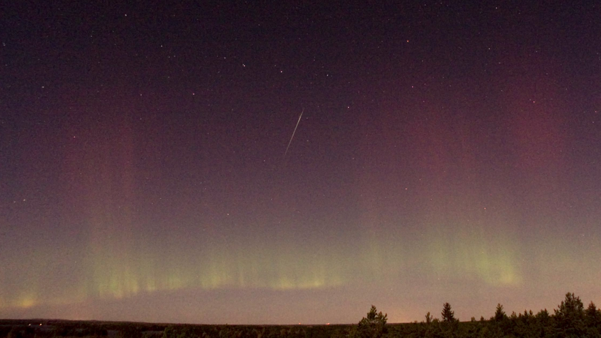 A lone shooting star streaks through a night sky riven with colorful aurora.